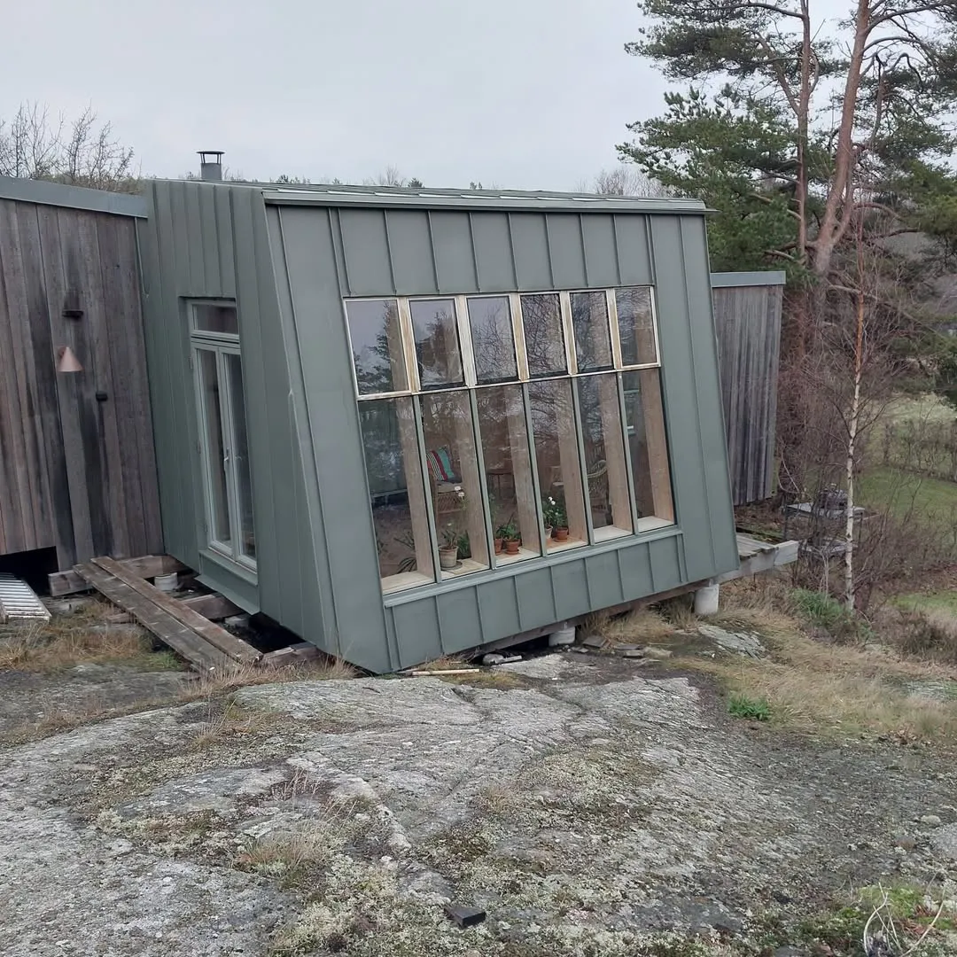 Grön plåtklädd tillbyggnad med stort fönsterparti, byggd på en berghäll bland tallar